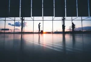 Silhuetas de viajantes em frente às janelas de vidro de um aeroporto durante o pôr do sol, com uma pessoa ao lado de uma mala de viagem enquanto utiliza o celular e o reflexo da luz no piso.