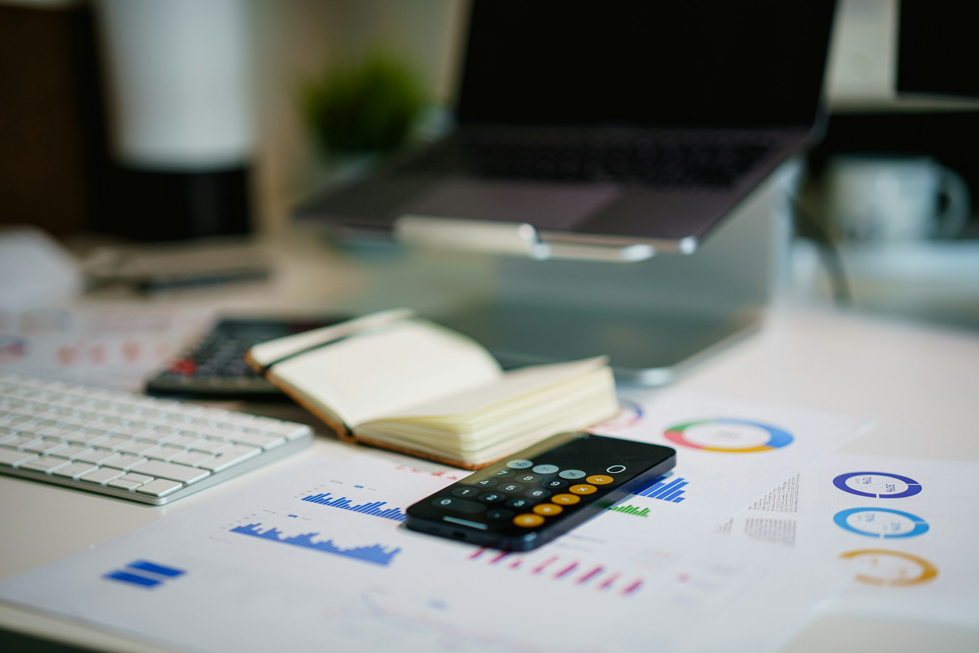 Mesa de trabalho com notebook, calculadora, teclado, caderno aberto e gráficos financeiros impressos sobre a mesa.