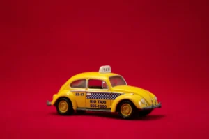 Miniatura de um táxi amarelo no estilo Volkswagen Fusca, com identificação "RIO TAXI", fotografada de perfil sobre fundo vermelho sólido.