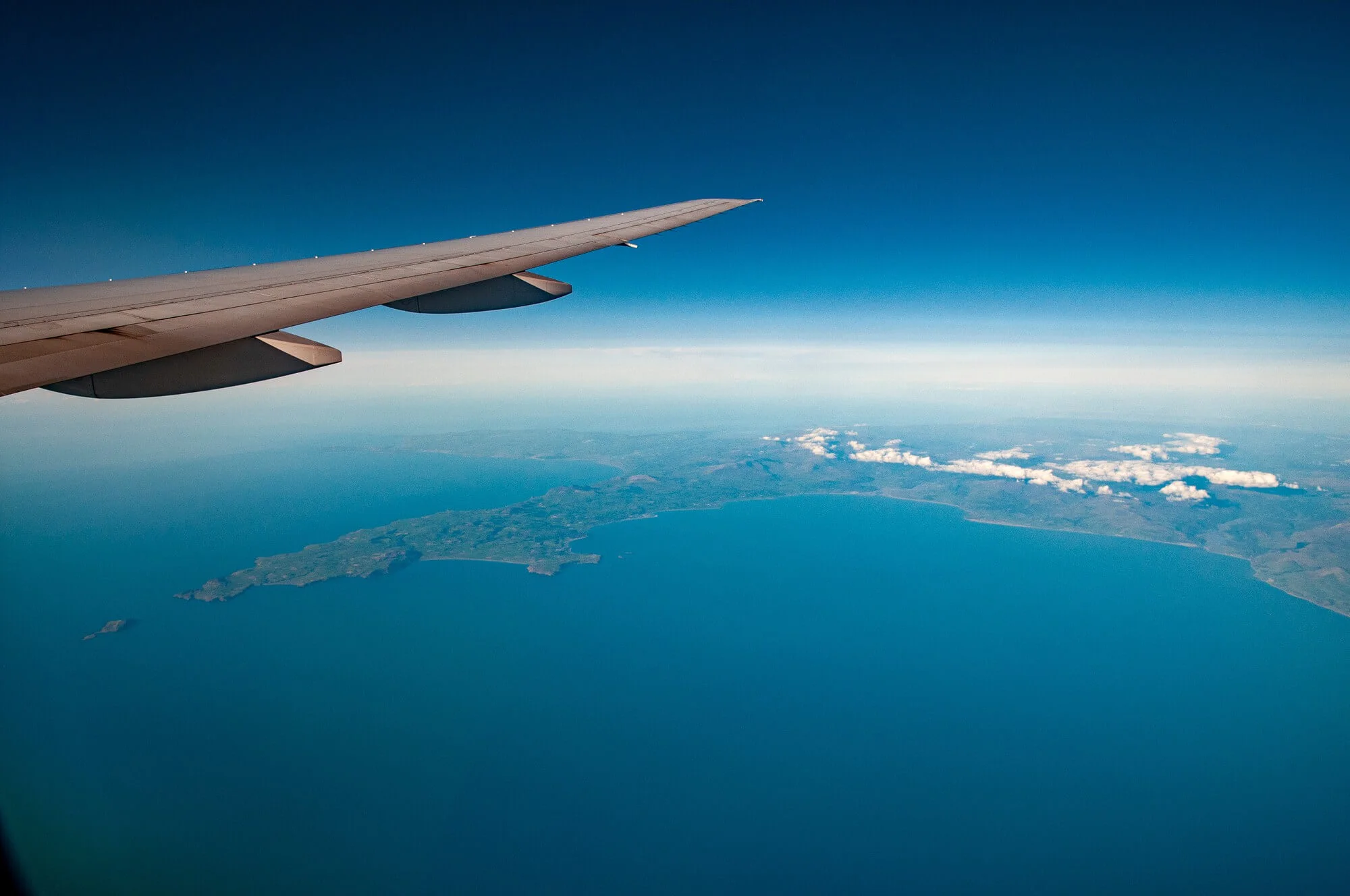 Uma vista aérea tirada da janela de um avião mostrando uma asa de avião branca e cinza no canto superior esquerdo. Abaixo da asa, grandes massas terrestres verdes e marrons são visíveis, cercadas pelo oceano azul. O céu acima é um azul profundo com uma pequena formação de nuvens brancas sobre as massas terrestres. O sol está brilhando do lado direito da imagem.