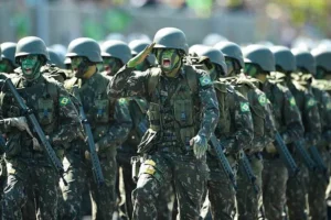 Fotografia de um pelotão de soldados do Exército Brasileiro marchando. Todos usam capacetes verdes, uniformes camuflados com a bandeira do Brasil na manga e pintura de camuflagem verde e preta no rosto. O soldado em destaque no centro está marchando, prestando continência com a mão direita no capacete e com a boca aberta em tom de marcha.